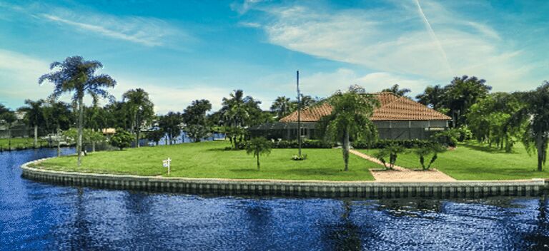 Waterfront residential property with seawall bordering a calm canal, featuring a large lawn, palm trees, and a house with a tile roof.