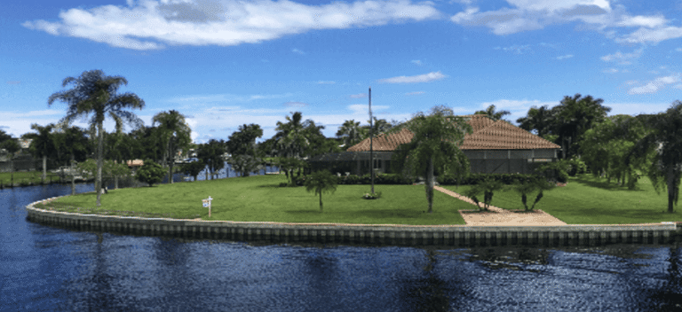 Waterfront residential property with seawall bordering a calm canal, featuring a large lawn, palm trees, and a house with a tile roof