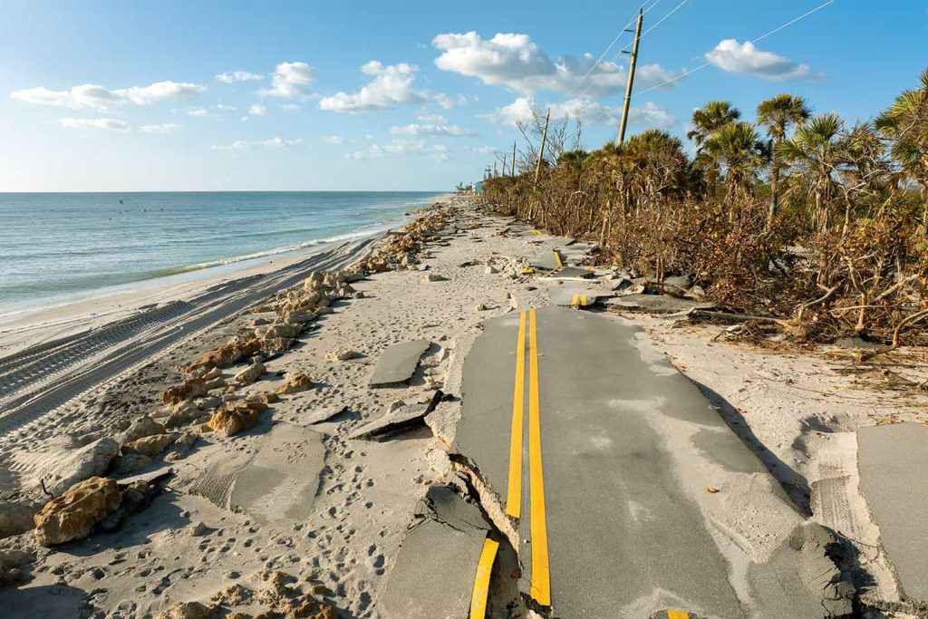 Damaged coastal road with crumbled asphalt and sand erosion caused by severe storm surge, running parallel to the beach and ocean.