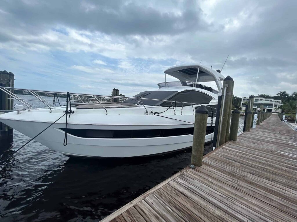 Luxury white yacht docked at a wooden marina pier on a cloudy day, with waterfront homes in the background.