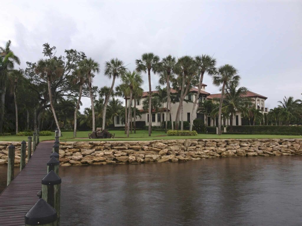 Waterfront estate with palm trees and a large stone seawall, viewed from a wooden dock on a calm waterway.