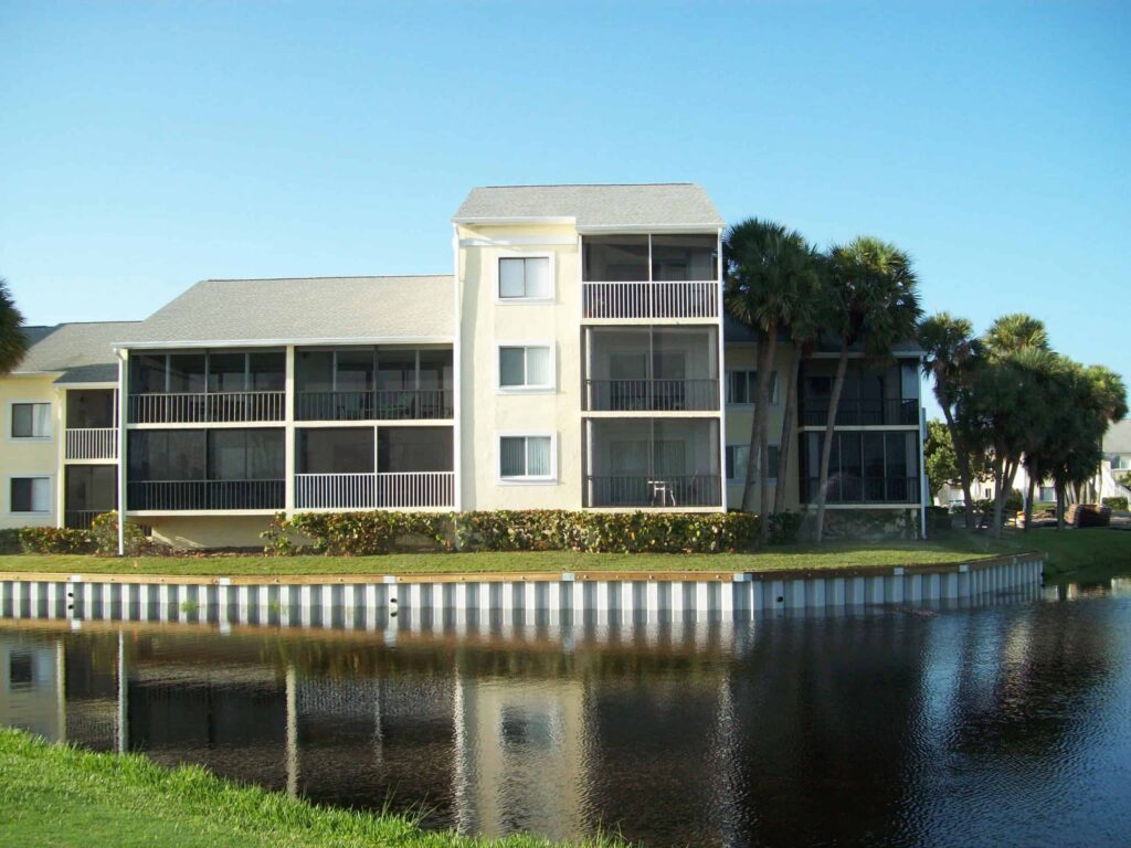 Three-story waterfront apartment building with screened balconies and metal seawall along calm pond