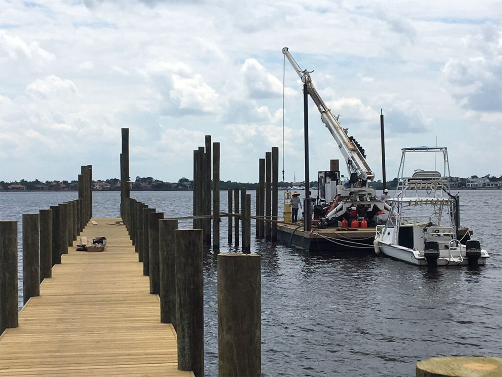 Marine construction crew working from a barge with a crane, installing or repairing wooden pilings on a long dock extending into the water.