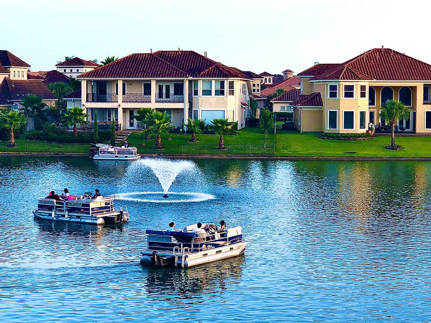 Pontoon boats cruising on a lake in front of large waterfront homes with a fountain in the center