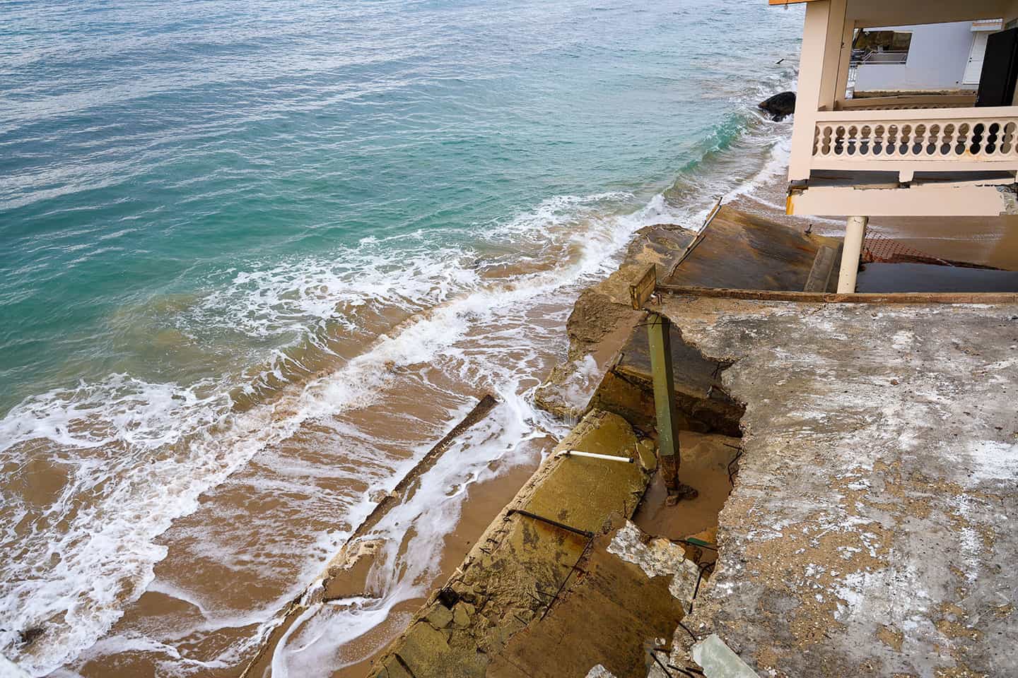 Coastal building with severe structural damage due to erosion and ocean waves, with collapsed concrete slabs on the shoreline.