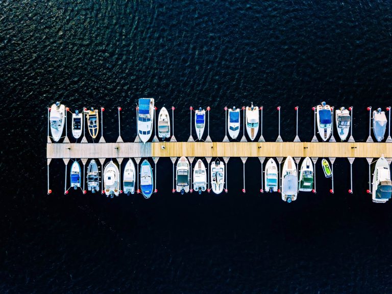 Aerial view of a marina dock with multiple boats moored on both sides in deep blue water.